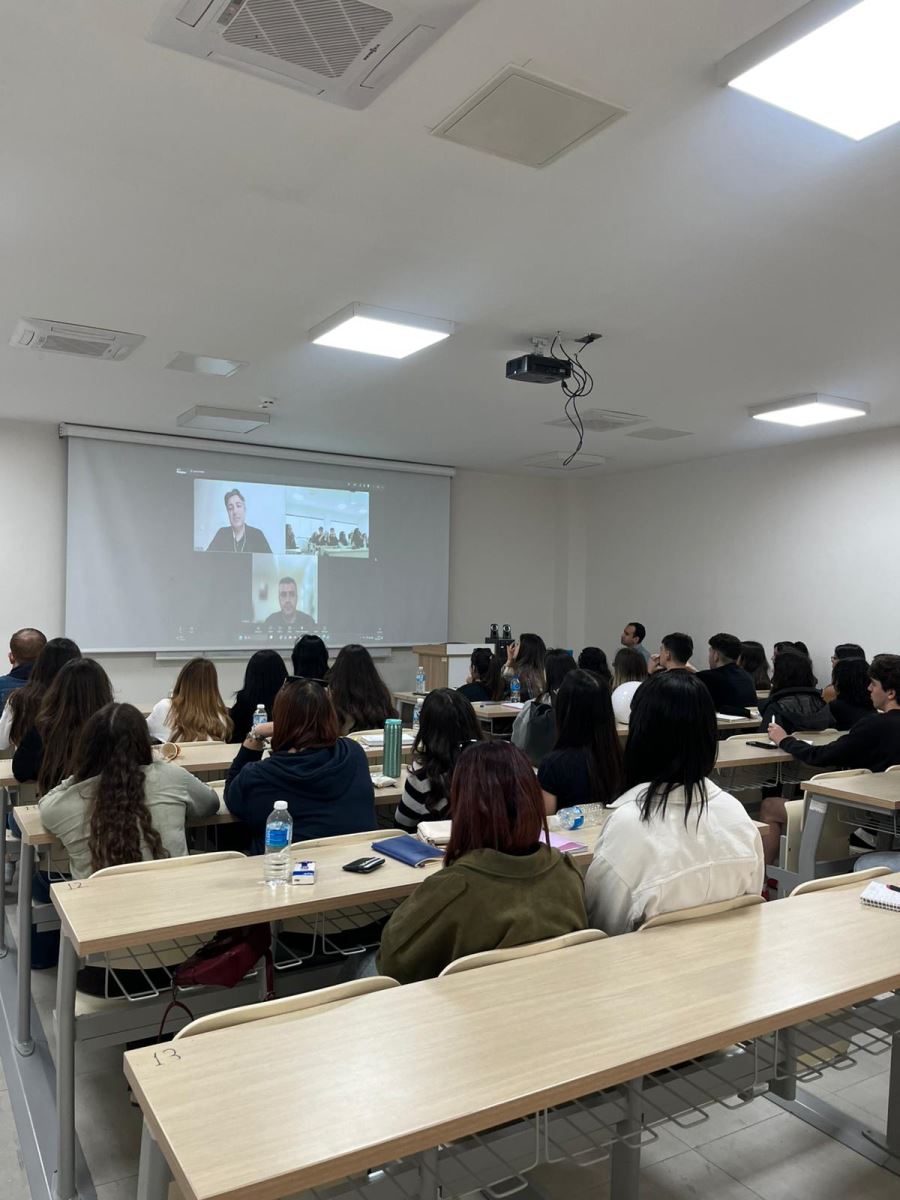 Sınıfın arka kısmından çekilmiş bu geniş açılı karede, dersliğin düzeni ve ortamın atmosferi ön planda. Öğrencilerin sıralarda düzenli bir şekilde oturdukları, ön taraftaki dev ekrana yansıtılan yayını dikkatle takip ettikleri görülüyor. Modern ve aydınlık bir sınıf ortamı göze çarpıyor.